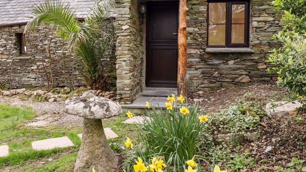 A stone house with a dark brown door and window, surrounded by a garden with yellow flowers and a mushroom-shaped stone. - Downrow Barn (Tintagel)