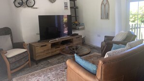 A living room with a television on a wooden stand, a bicycle on the wall, and a window with a view of greenery. - Downrow Barn (Tintagel)