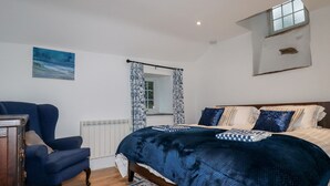 A bedroom with a bed, a chair, a radiator, a window with curtains, and a painting on the wall. - Downrow Barn (Tintagel)
