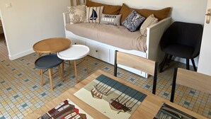Un salon chaleureux avec un canapé, deux petites tables d’appoint, un fauteuil noir et un luminaire suspendu en fibres tressées.