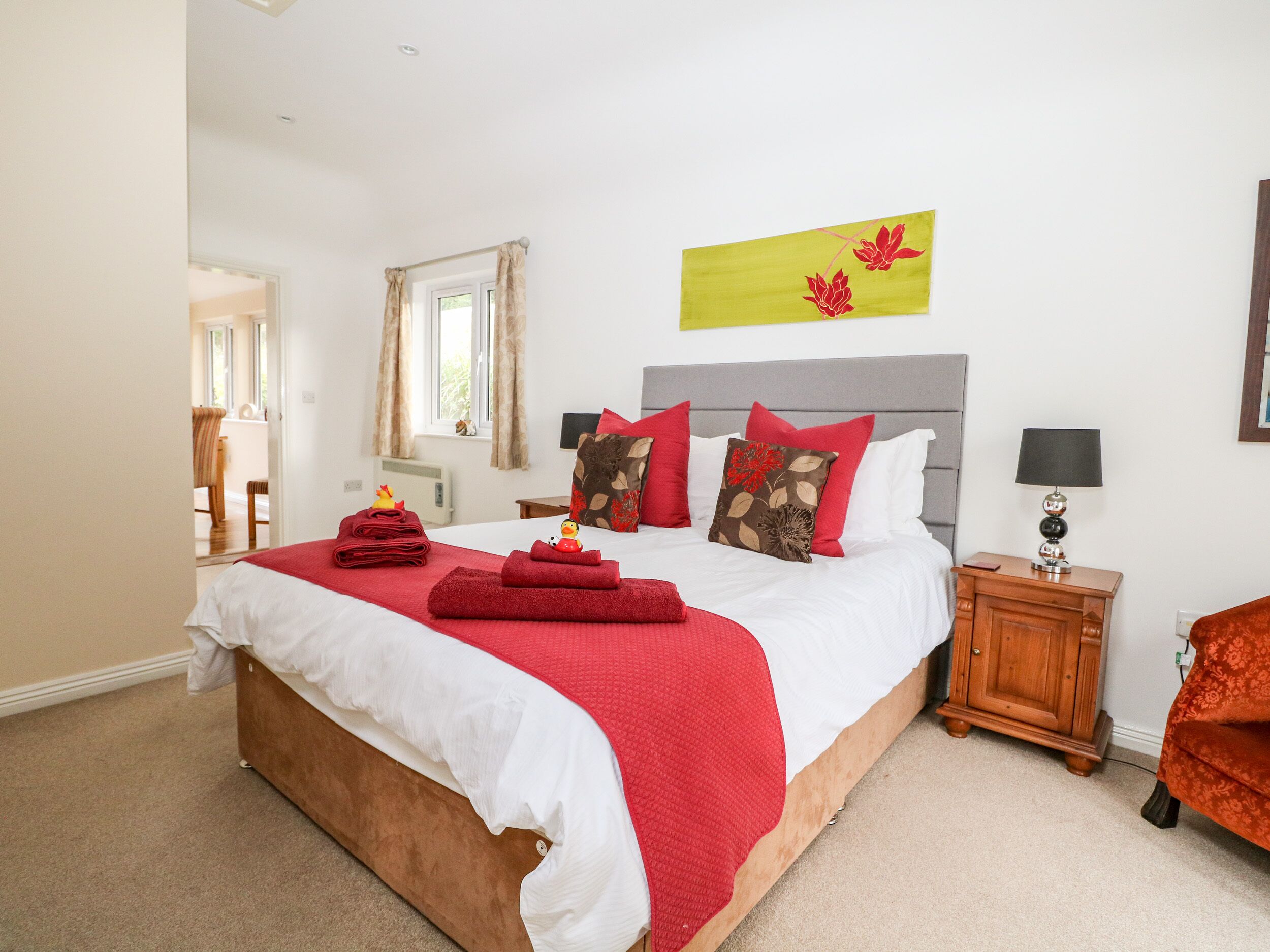 A neatly made bed with red and white bedding, a wooden nightstand with a lamp, and a window with curtains.