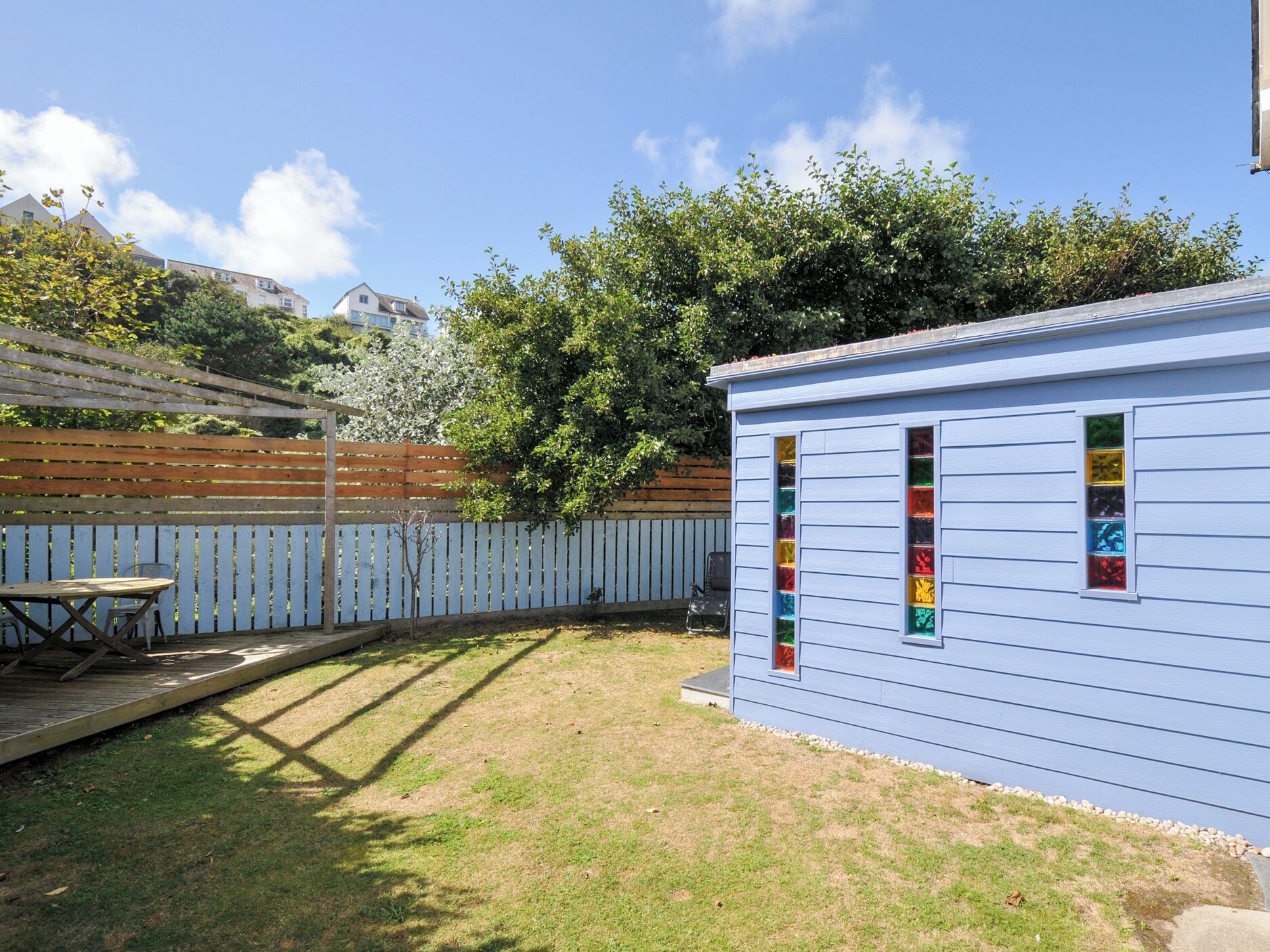Un abri de jardin bleu avec des fenêtres colorées, une terrasse en bois et une cour arrière verdoyante.