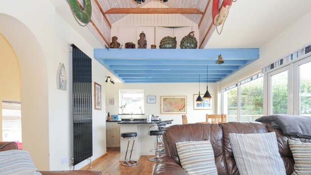 A cozy living room with a brown leather sofa, a kitchen area with blue accents, and a dining area with a wooden table and chairs. - Beachside (Wadebridge)