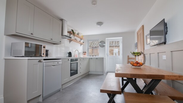 A modern kitchen with grey cabinets, a microwave, and a sink. There is a dining table with a fruit bowl and a TV mounted on the wall. - Fell Briggs Cottage (Redcar)