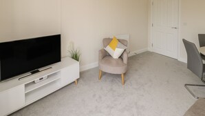 A modern living room with a flat-screen TV on a white entertainment unit, a beige armchair with a yellow pillow, a small plant, and a dining area with a chair and table. - Valley View (Whitby)