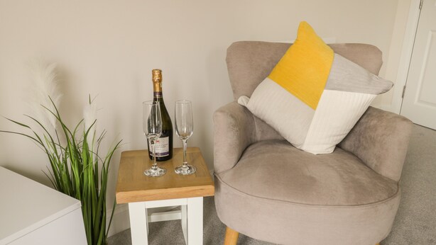 A beige armchair with a yellow pillow, a small wooden table with a bottle of wine and two glasses, and a potted plant. - Valley View (Whitby)