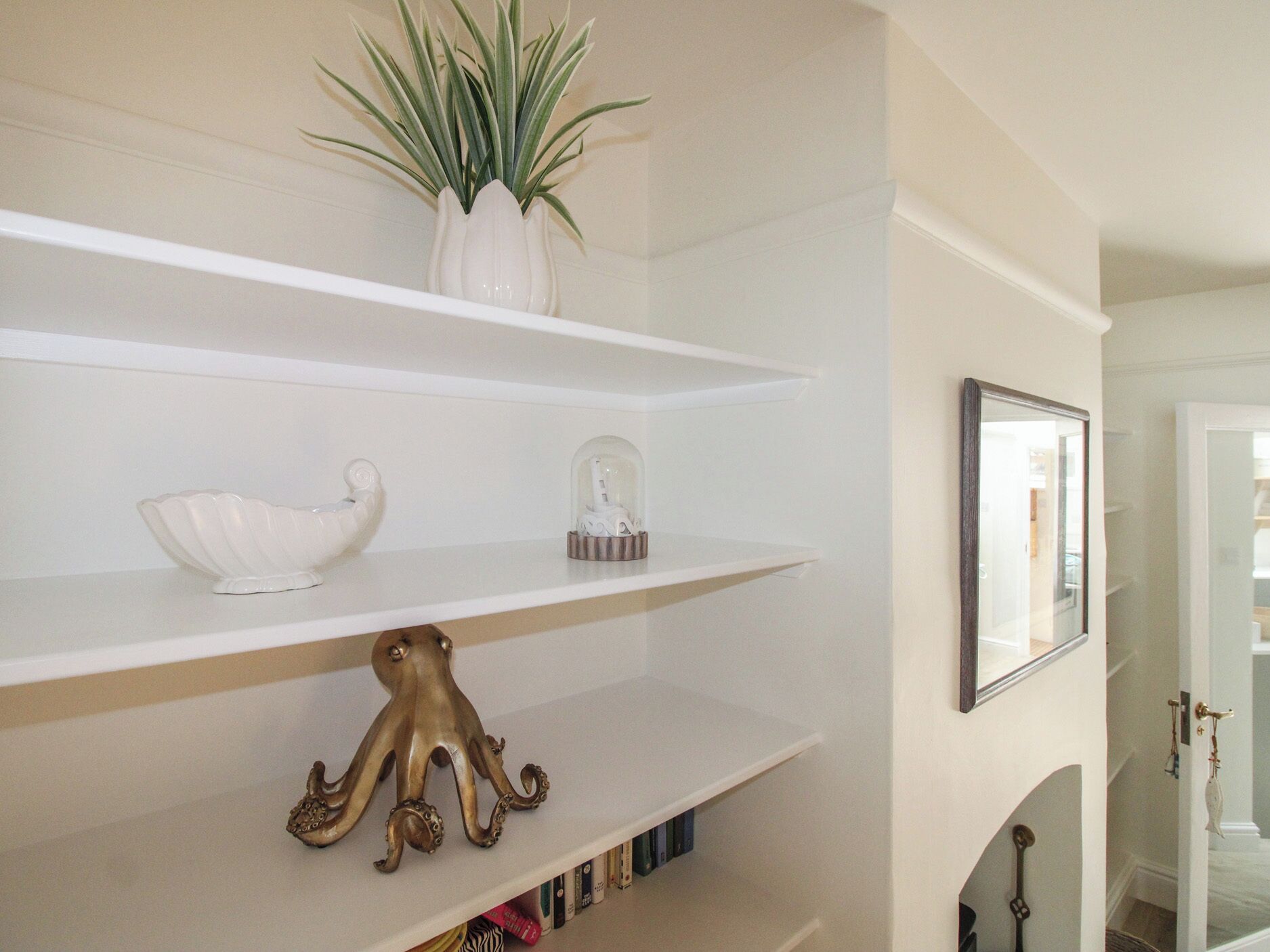Una habitación con estantes blancos, una planta en maceta, un objeto decorativo de vidrio y un cuadro en la pared.