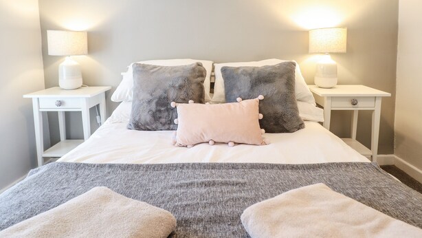 A neatly arranged bedroom with a bed, two bedside tables, and a wall-mounted shelf with framed pictures. - Driftwood Terrace (Porthmadog)