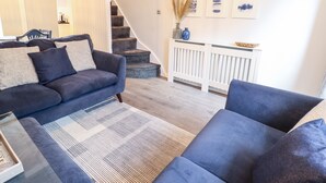 A modern living room with a dark blue sofa, a glass coffee table, and a patterned rug. A staircase leads to an upper level. - Driftwood Terrace (Porthmadog)