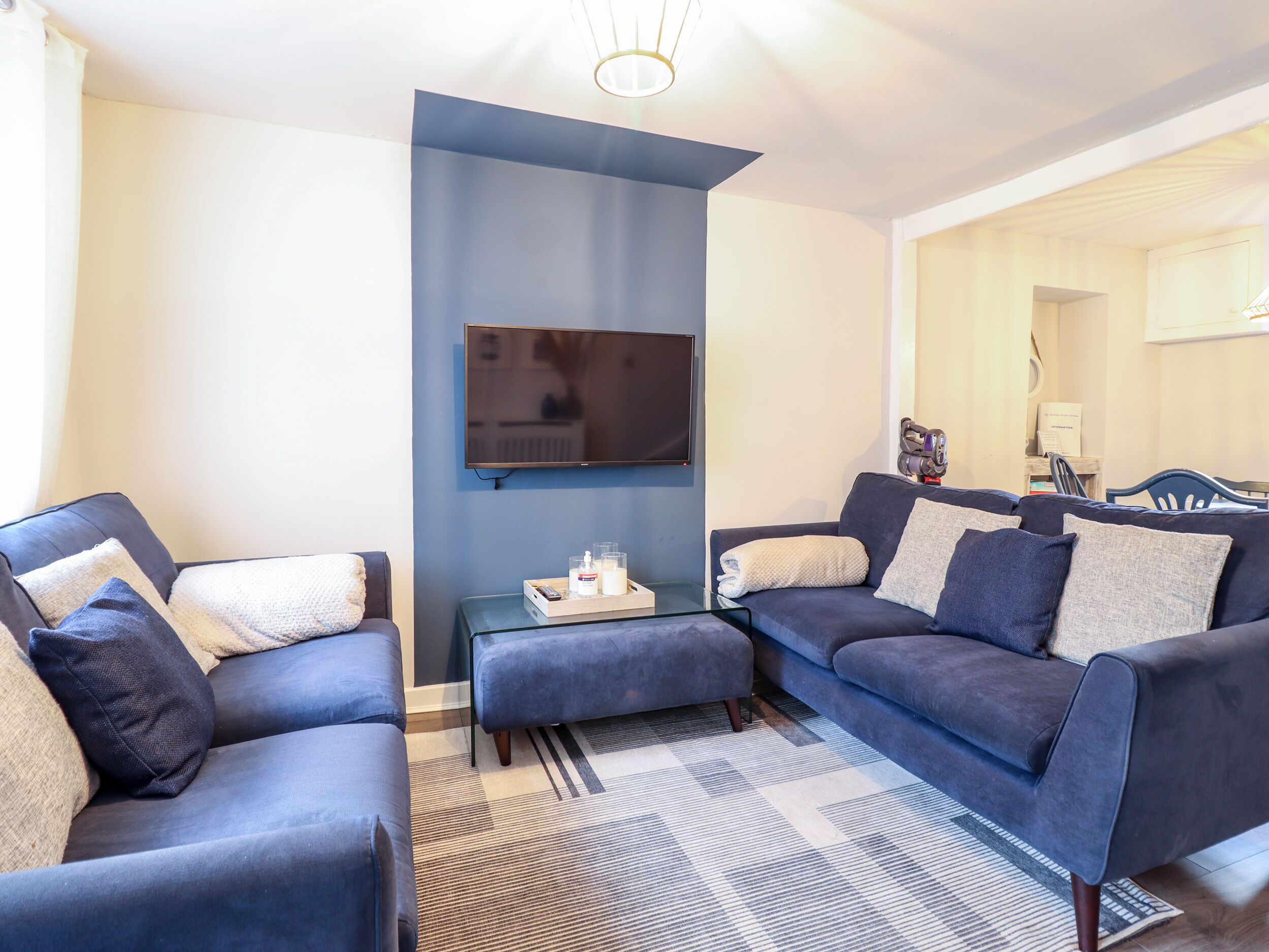 A modern living room with a large flat-screen TV mounted on the wall, a sofa set, a coffee table, and a patterned rug.