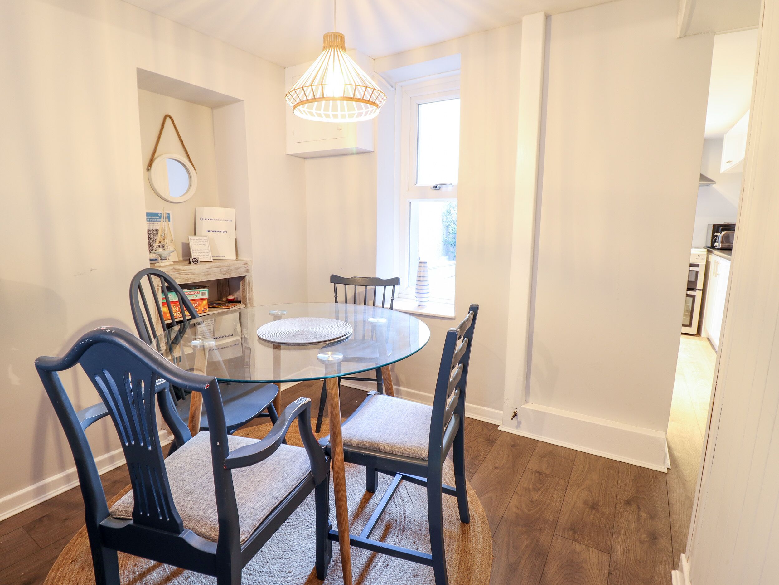 A dining area with a round glass table, four chairs, a hanging light fixture, and a window with curtains.