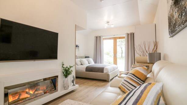 A modern living room with a fireplace, a sofa, a television, and a bed in the background. - Westfields (Launceston)
