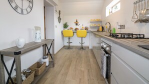 A modern kitchen with a large wall clock, two yellow bar stools, a wooden table with baskets, and a stainless steel range hood. - Westfields (Launceston)