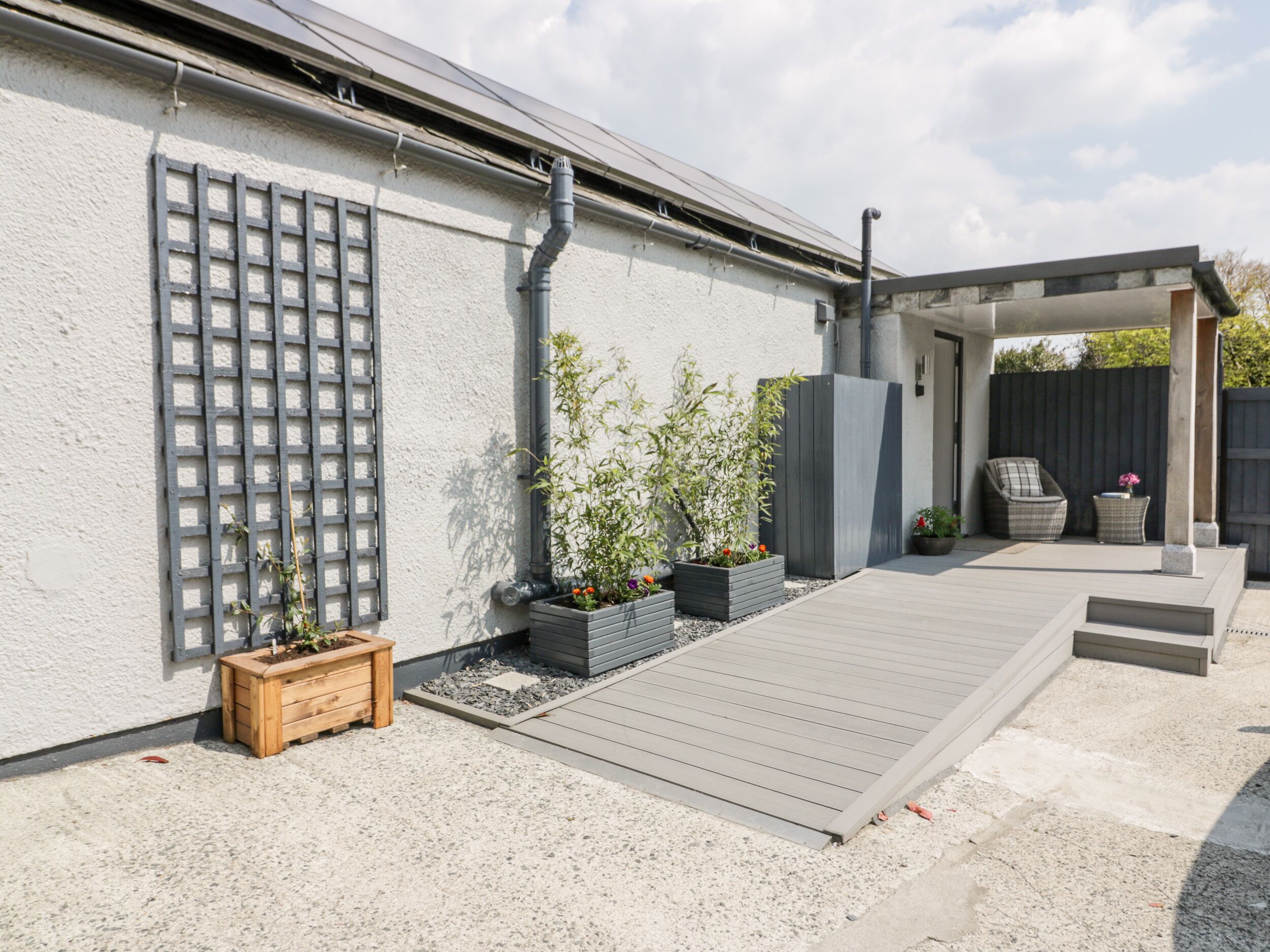 Un patio con cubierta de madera, macetas con plantas, una pérgola y una zona de estar con una silla y una mesa.