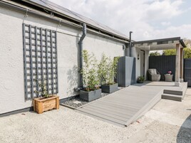 Un patio con cubierta de madera, macetas con plantas, una pérgola y una zona de estar con una silla y una mesa.