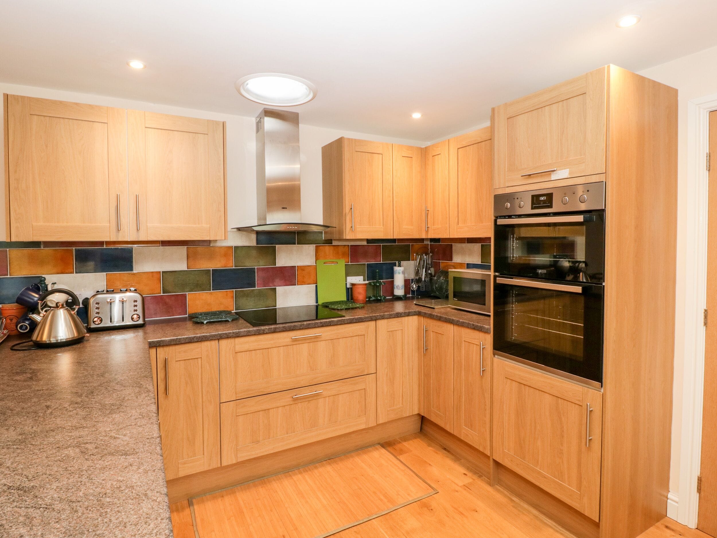 A modern kitchen with wooden cabinets, a stainless steel range hood, a double oven, and a colorful backsplash.