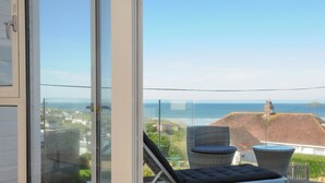 Un balcon doté d’une porte vitrée, de deux fauteuils en osier, d’un banc rembourré et offrant une vue sur la mer et les bâtiments.
