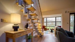 A modern interior with a wooden desk, a lamp, a potted plant, a grey sofa, and a staircase with exposed pipes. - Riverside Park 5 (Keswick)