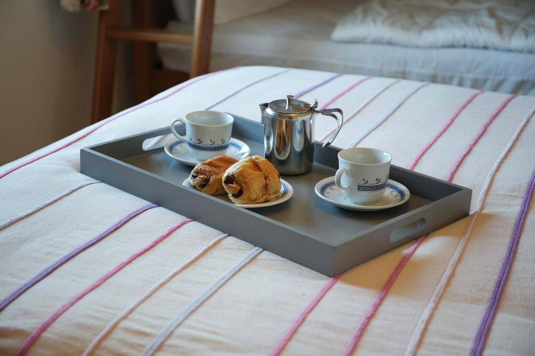 A tray with croissants, a teapot, and two cups on a bed with striped bedding.