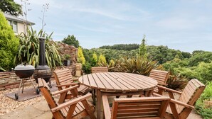 An outdoor wooden table and chairs set on a patio with a stone floor, surrounded by various plants and a stone wall. - The Stable (Lydney)