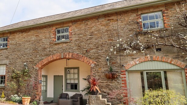 A stone building with a brick archway entrance, a small porch with wicker furniture, and a large window on the right. - The Stable (Lydney)