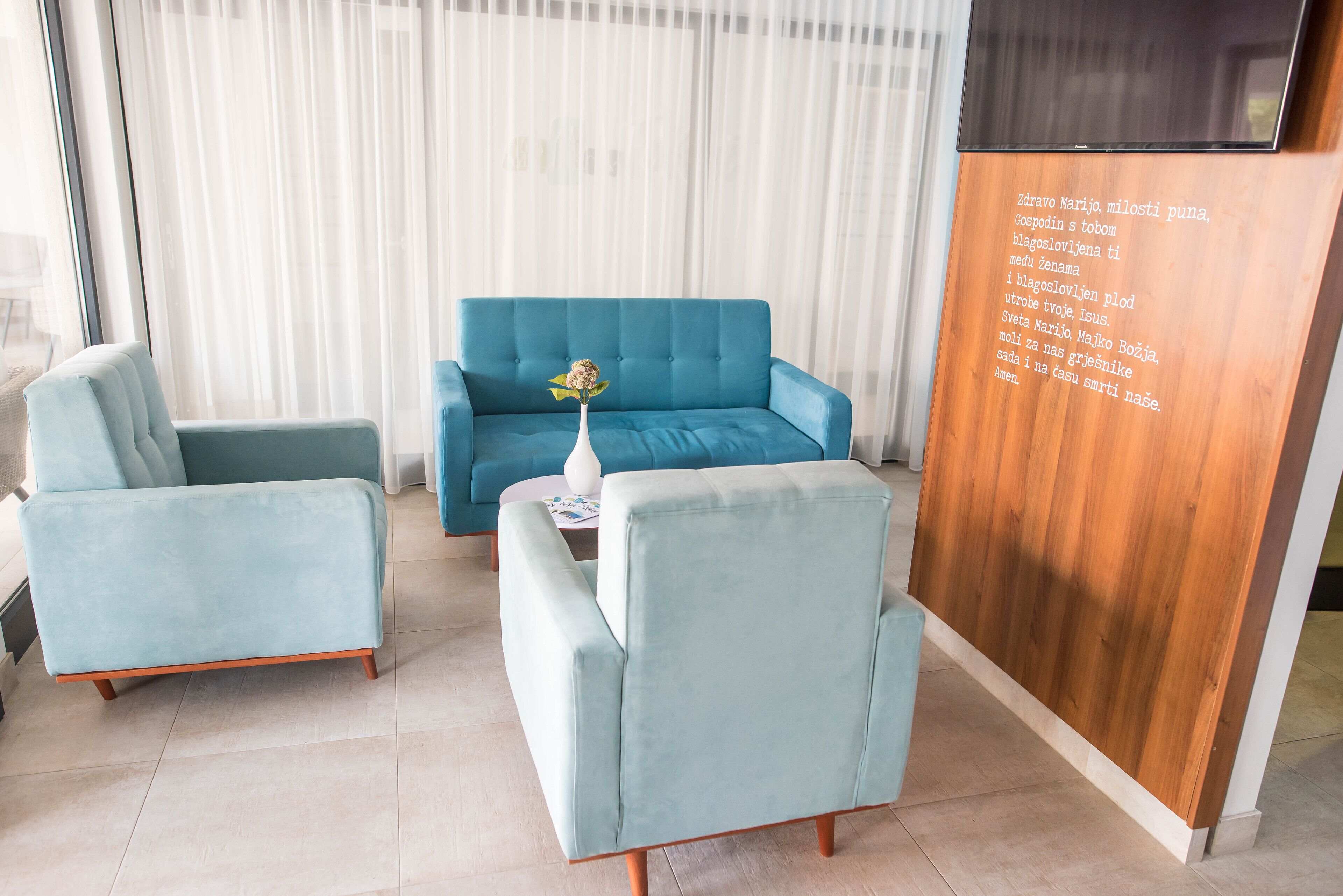 A modern living room with a blue sofa, two matching armchairs, and a small side table with a vase of flowers. A wooden panel with text is visible on the right.