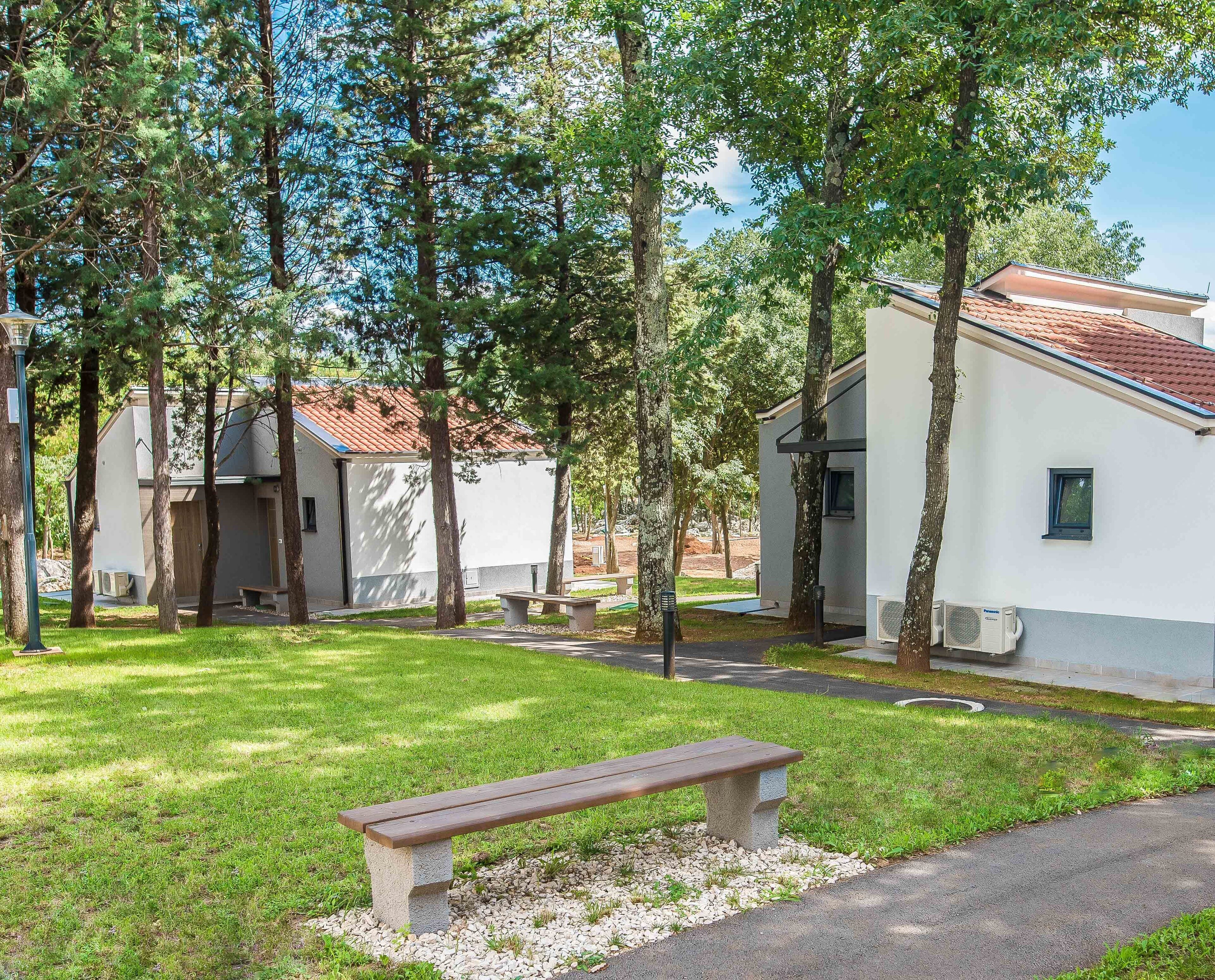 A serene outdoor area with a wooden bench, a pathway, and a row of white buildings with red-tiled roofs.
