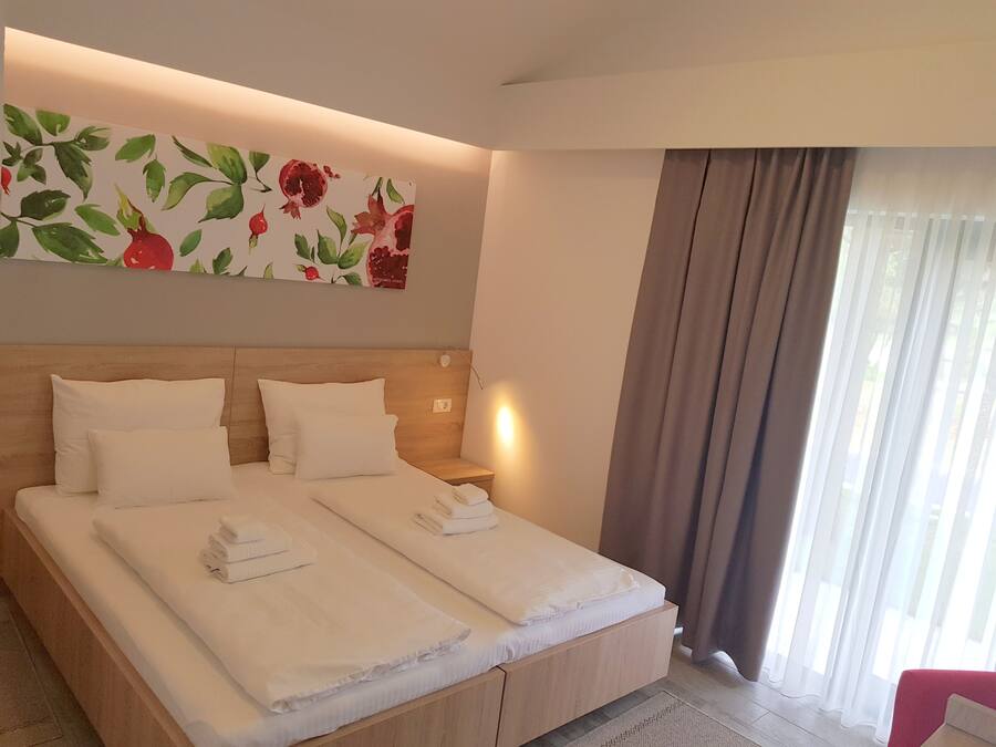 A neatly made double bed with white linens and towels, a wooden headboard, a wall-mounted artwork of red flowers, a bedside table with a lamp, and a window with curtains.