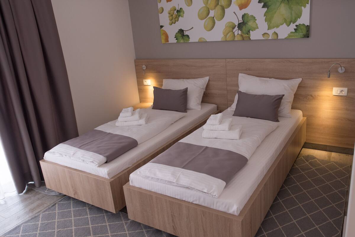 A hotel room with two beds, each with a wooden headboard, white linens, and a brown pillow. There is a wall-mounted light above each bed and a painting of grapes on the wall.