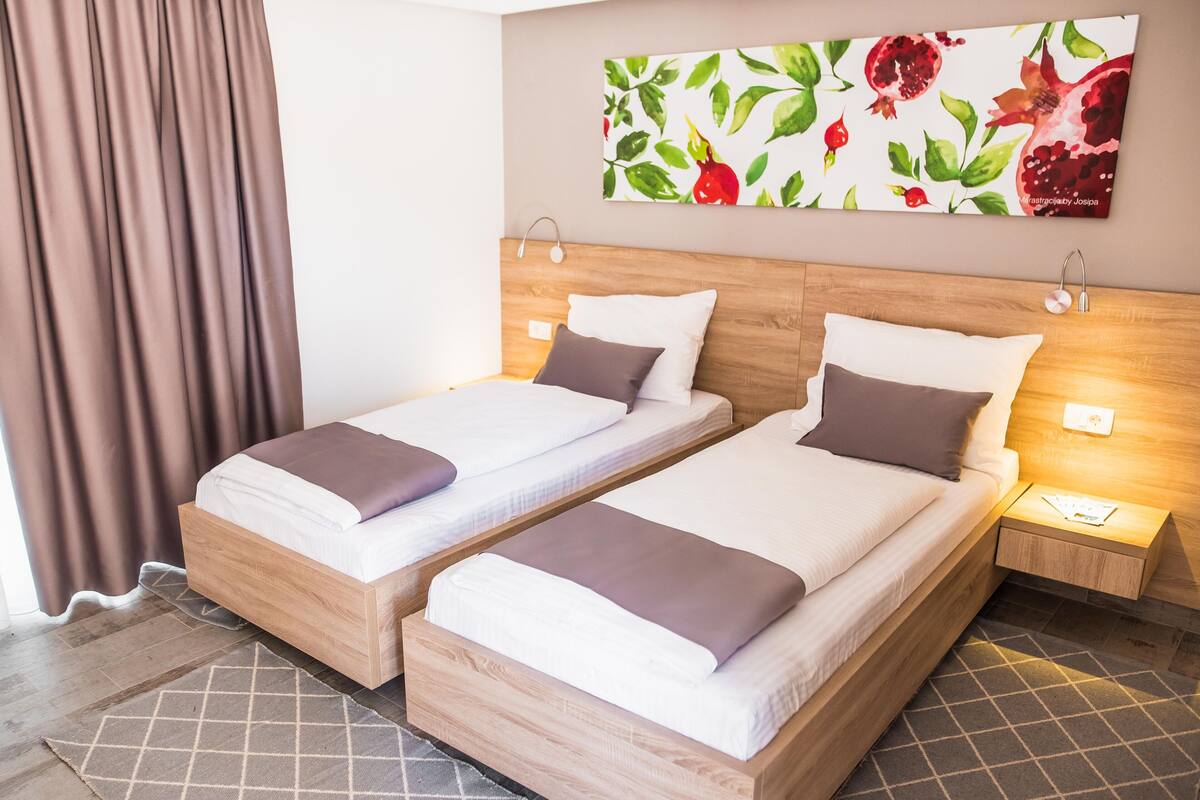 A hotel room with two single beds, a wooden headboard, a nightstand with a lamp, and a pomegranate-themed artwork on the wall.