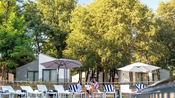 Zona de piscina con sillones reclinables y sombrillas, rodeada de árboles y un edificio.