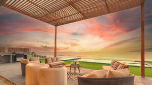 A covered outdoor seating area with a view of the ocean at sunset, featuring wicker furniture and a poolside bar.