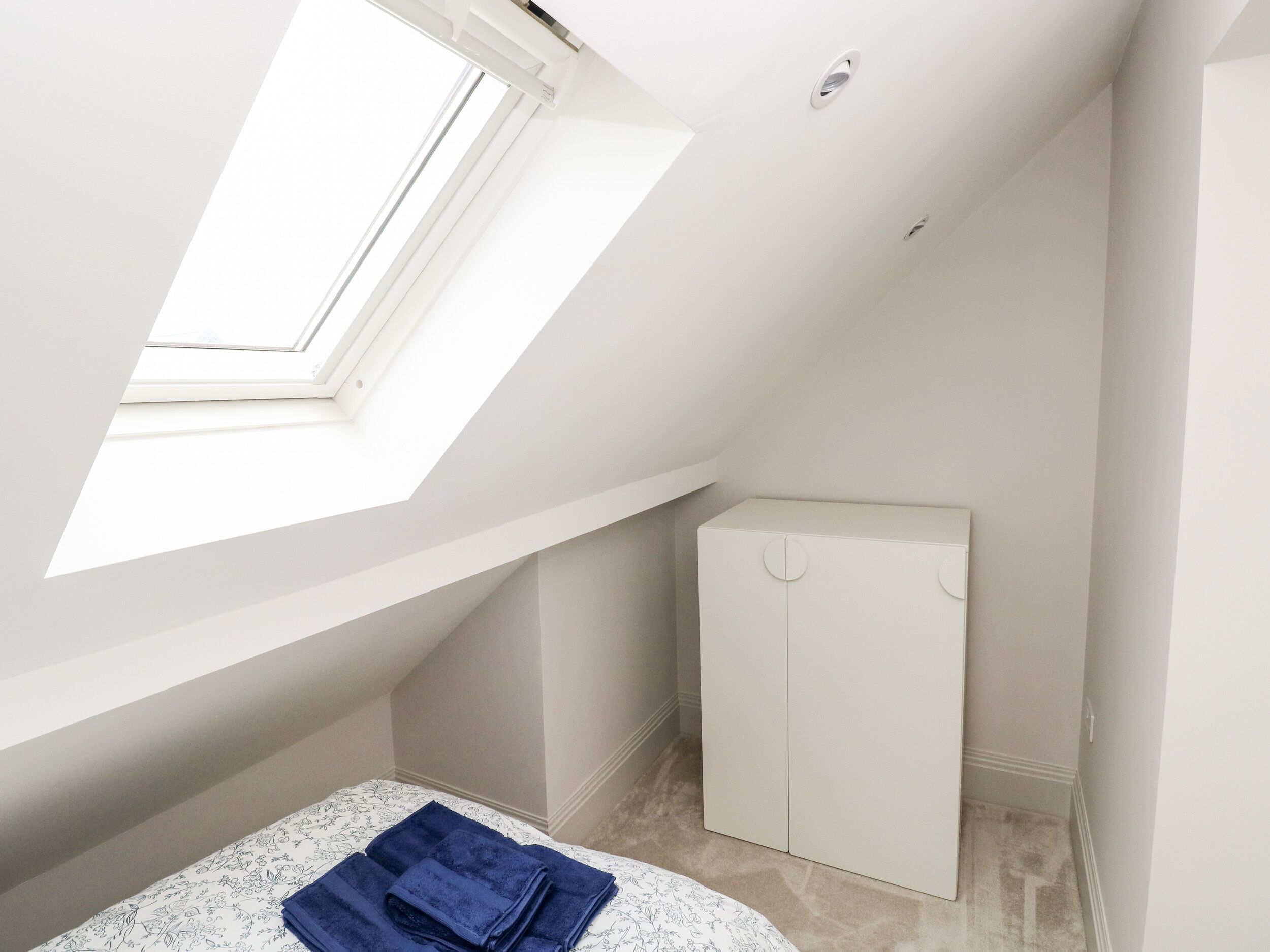 Una habitación pequeña y blanca con techo inclinado, tragaluz y armario.