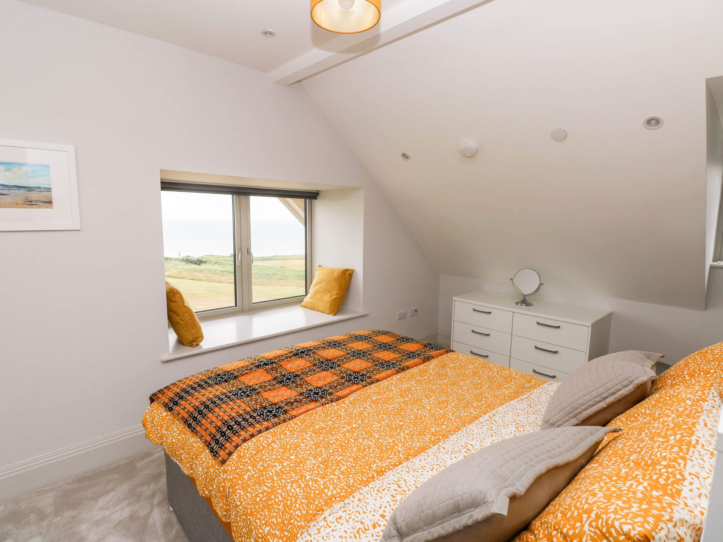 Une chambre avec une grande fenêtre, un lit recouvert d’une couvre-lit à motifs orange et blanc, une commode blanche et un petit miroir rond.