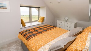 Un dormitorio con un gran ventanal, una cama con colcha naranja y blanca, un tocador blanco y un espejo redondo pequeño.
