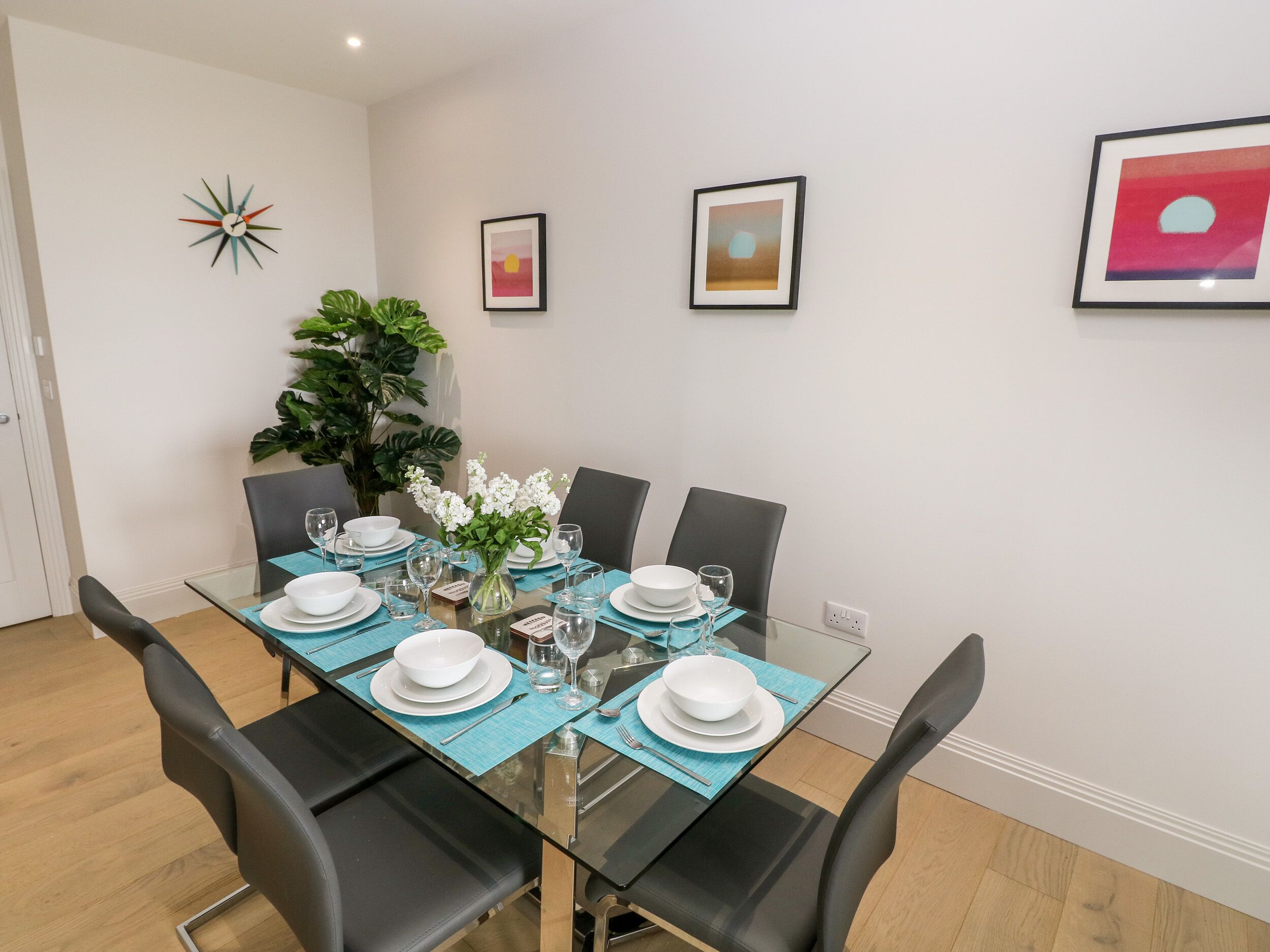 A dining area with a glass table set for six, surrounded by black chairs. There are framed artworks on the walls and a potted plant on the table.