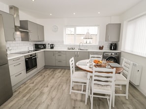 A modern kitchen with white cabinets, a built-in oven, and a dining area with a round table and white chairs. - Trigfa (Moelfre)