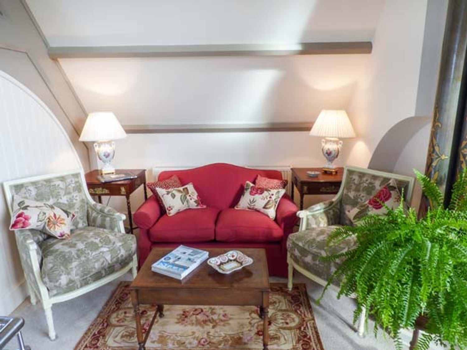 A living room with a red sofa, floral armchairs, a coffee table, and two lamps.