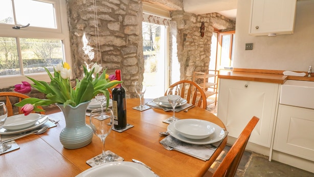 A dining area with a stone wall, wooden table, and white plates. - The Priory (Bakewell)