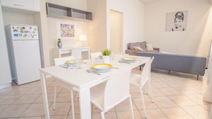 Una cocina moderna con armarios blancos, una mesa de comedor para dos personas y un sofá gris al fondo.