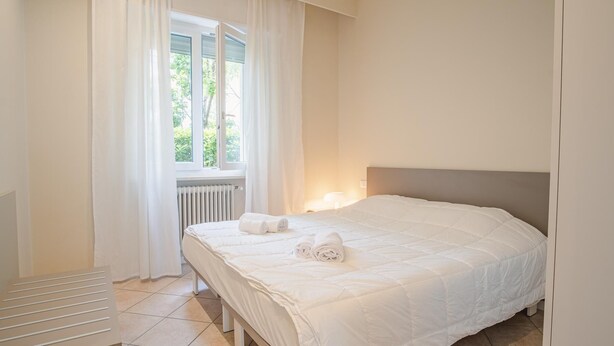 Una cama bien tendida con sábanas blancas y toallas enrolladas, una ventana con cortinas blancas y un radiador debajo de la ventana.