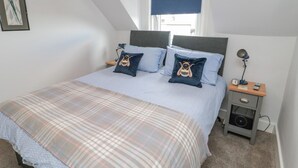 A bedroom with a bed, two pillows, a nightstand, a lamp, a clock, and a small drawer unit. - 3 St. Cuthberts Garth (Bamburgh)
