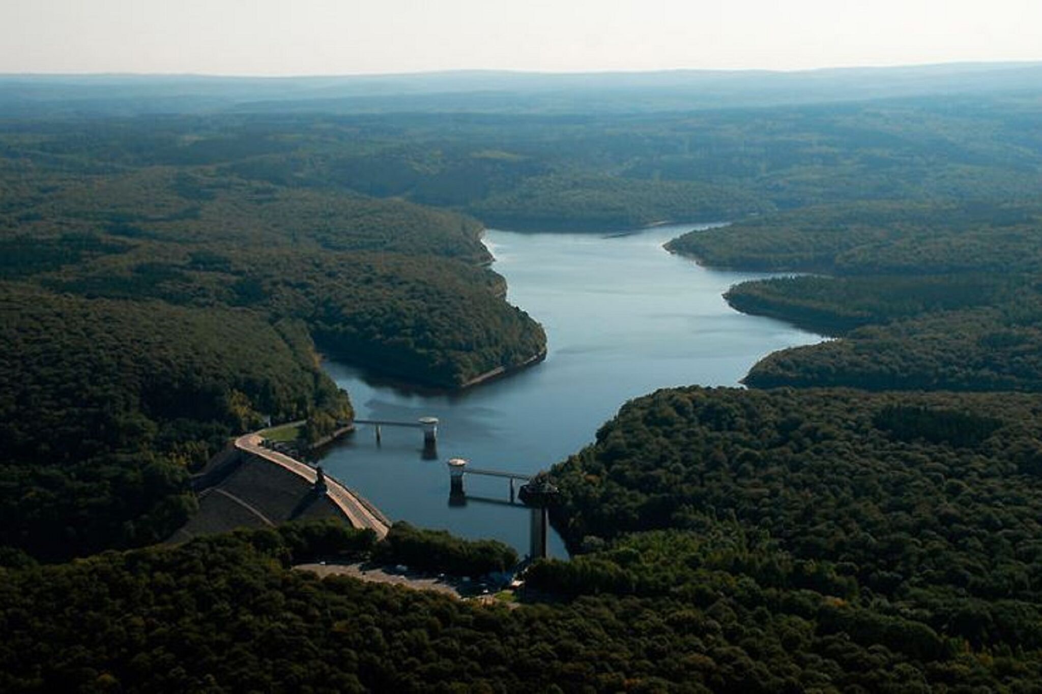 Un barrage surmonté d’un pont et un réservoir d’eau, le tout entouré de collines boisées.
