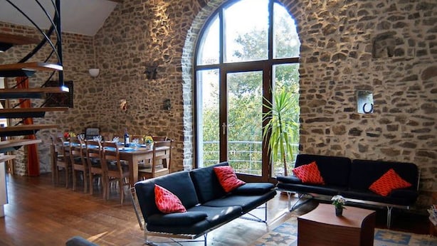 A spacious living area with a stone wall, a large arched window, and a dining area with wooden furniture. - Holiday Home in Verviers with Sauna & Fireplace (Ensival)