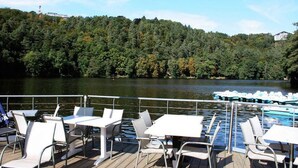 A deck with tables and chairs overlooking a lake, surrounded by dense forest. - Holiday Home in Verviers with Sauna & Fireplace (Ensival)