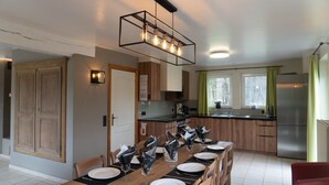 A dining area with a long wooden table set for six, a kitchen with wooden cabinets, a stainless steel refrigerator, and a hanging light fixture. - Holiday Home in Stoumont near Coo Waterfalls (Rahier)