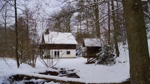 Eine rustikale Holzhütte mit schneebedecktem Dach, umgeben von Bäumen und einem kleinen Bach.