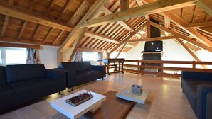 Un vaste espace de vie de style loft, doté de hauts plafonds et de poutres apparentes. On y trouve deux canapés sombres et une table basse agrémentée d’objets décoratifs. Un téléviseur est fixé au mur, et une cheminée est visible au fond.