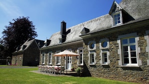 A stone building with a patio area featuring tables and chairs, a large umbrella, and a well-maintained lawn. - Le Hameau du Château – Elegance and serenity (Libin)