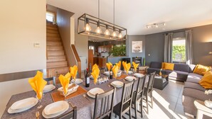 Un comedor con una mesa larga dispuesta para seis personas, una escalera, un sofá y una cocina al fondo.